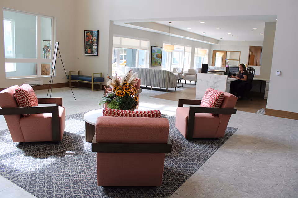 A bright and spacious common area in a senior living facility featuring four pink armchairs arranged around a circular table with a floral centerpiece. In the background, there is a reception desk with a staff member working, additional seating areas with striped and beige chairs, large windows letting in natural light, and artwork on the walls.