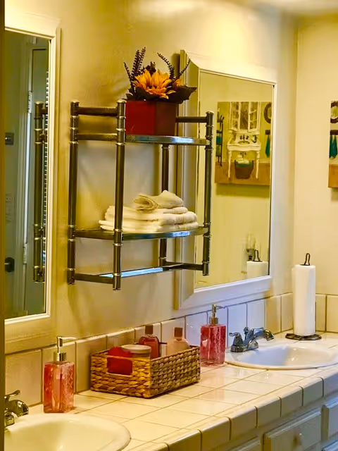 A bathroom countertop with two sinks, each with a silver faucet. On the countertop, there are two pink liquid soap dispensers, a wicker basket holding toiletries, and a paper towel holder with a roll of paper towels. Above the countertop is a large mirror and a metal wall shelf holding folded white towels and a red pot with a sunflower and other decorative plants. A painting of a bathroom scene is visible on the wall next to the mirror.