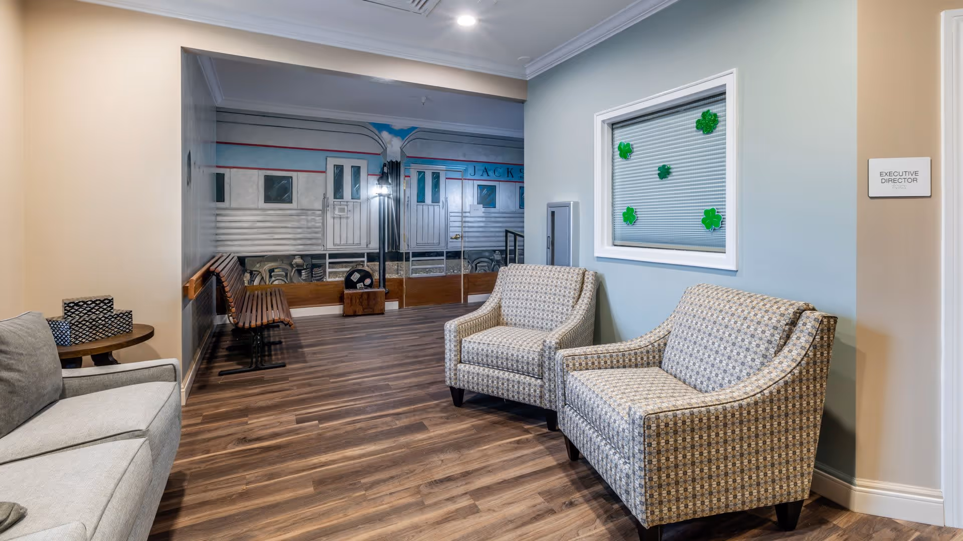A waiting area with two patterned armchairs and a gray sofa. There is a small round table with tissue boxes on it. The floor is wooden, and the walls are painted in light beige and blue. A window with green shamrock decorations and a sign labeled 'Executive Director' are visible. The back wall features a mural of a vintage train car and a wooden bench.
