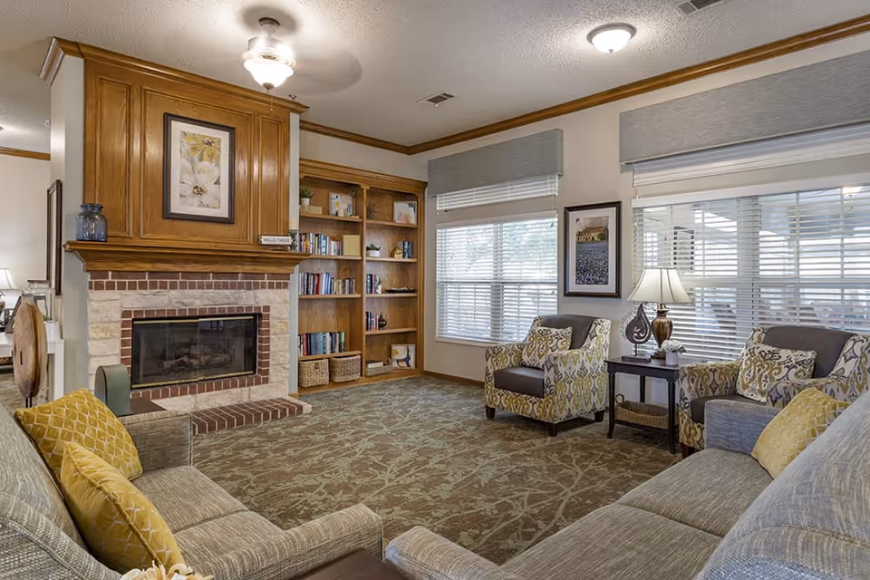 A cozy living room with a stone and wood fireplace, built-in wooden bookshelves filled with books and decorative items, two patterned armchairs with cushions, a sofa with yellow pillows, a side table with a lamp, and large windows with blinds letting in natural light.