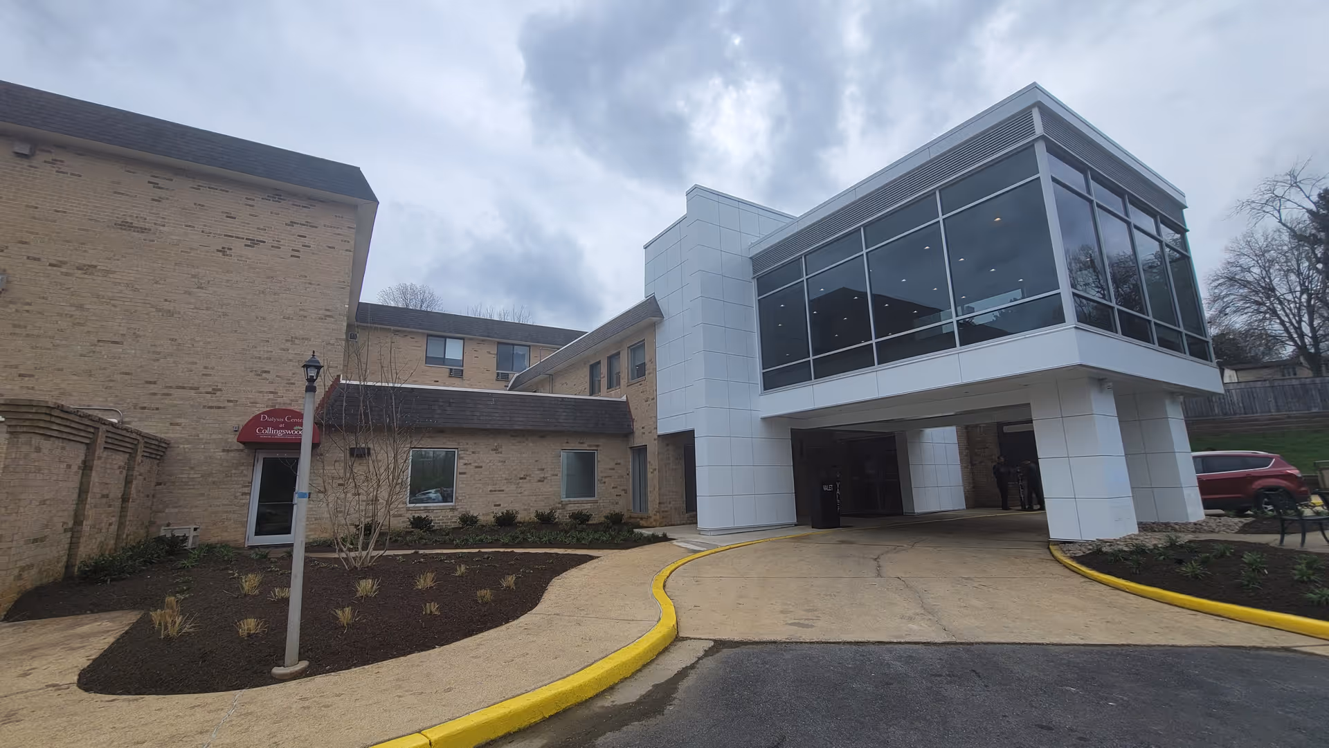 Exterior view of Collingswood Rehabilitation & Healthcare Center showing a two-story building with beige brick walls and a modern white structure with large glass windows extending over the driveway. The driveway curves with a yellow curb, and there are landscaped areas with small plants and a lamppost near the entrance.