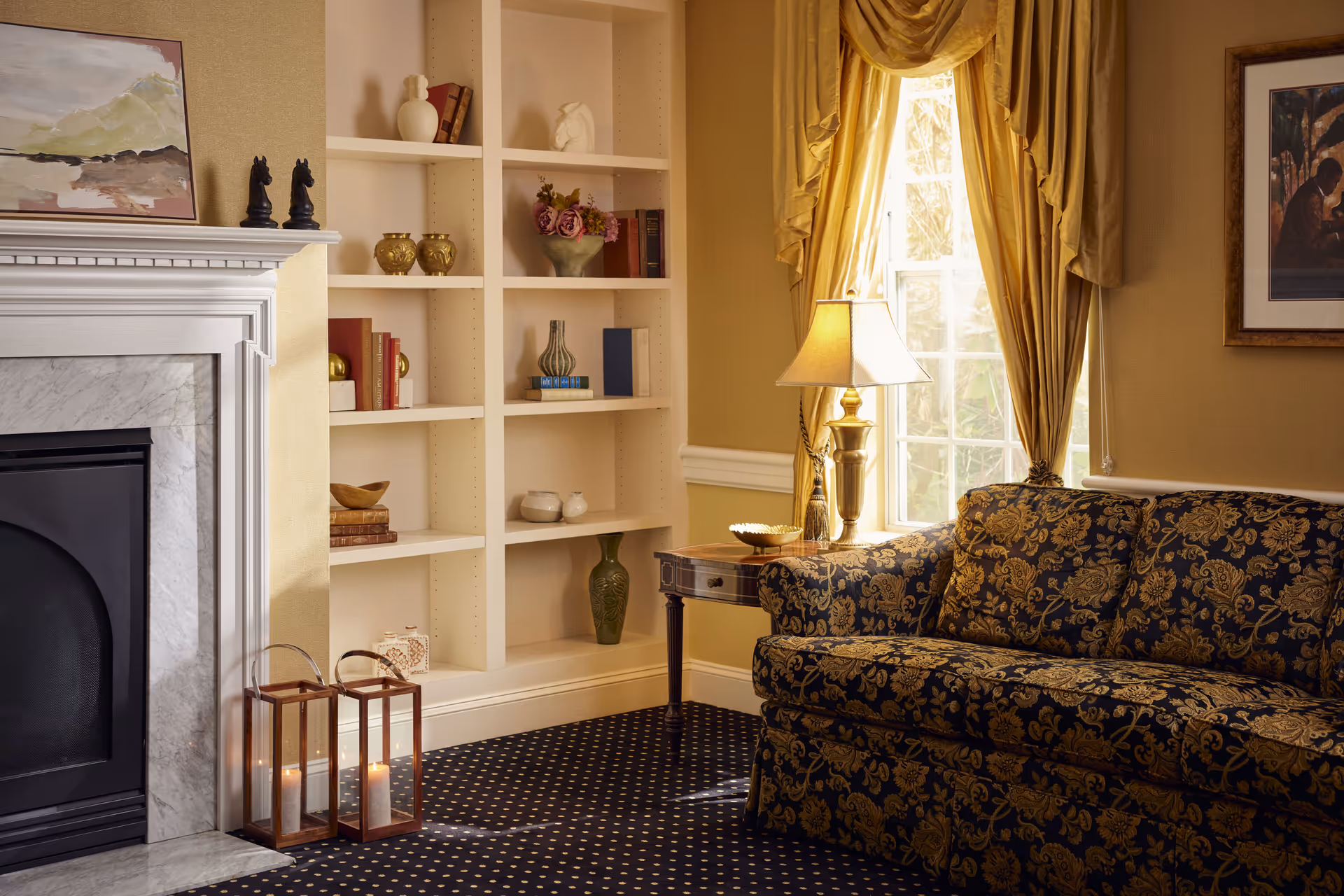 A cozy living room corner featuring a patterned dark blue and gold sofa, a wooden side table with a lamp, a window with gold curtains, a white built-in bookshelf with decorative items and books, a white marble fireplace with two lit lantern candles on the floor, and framed artwork on the walls.