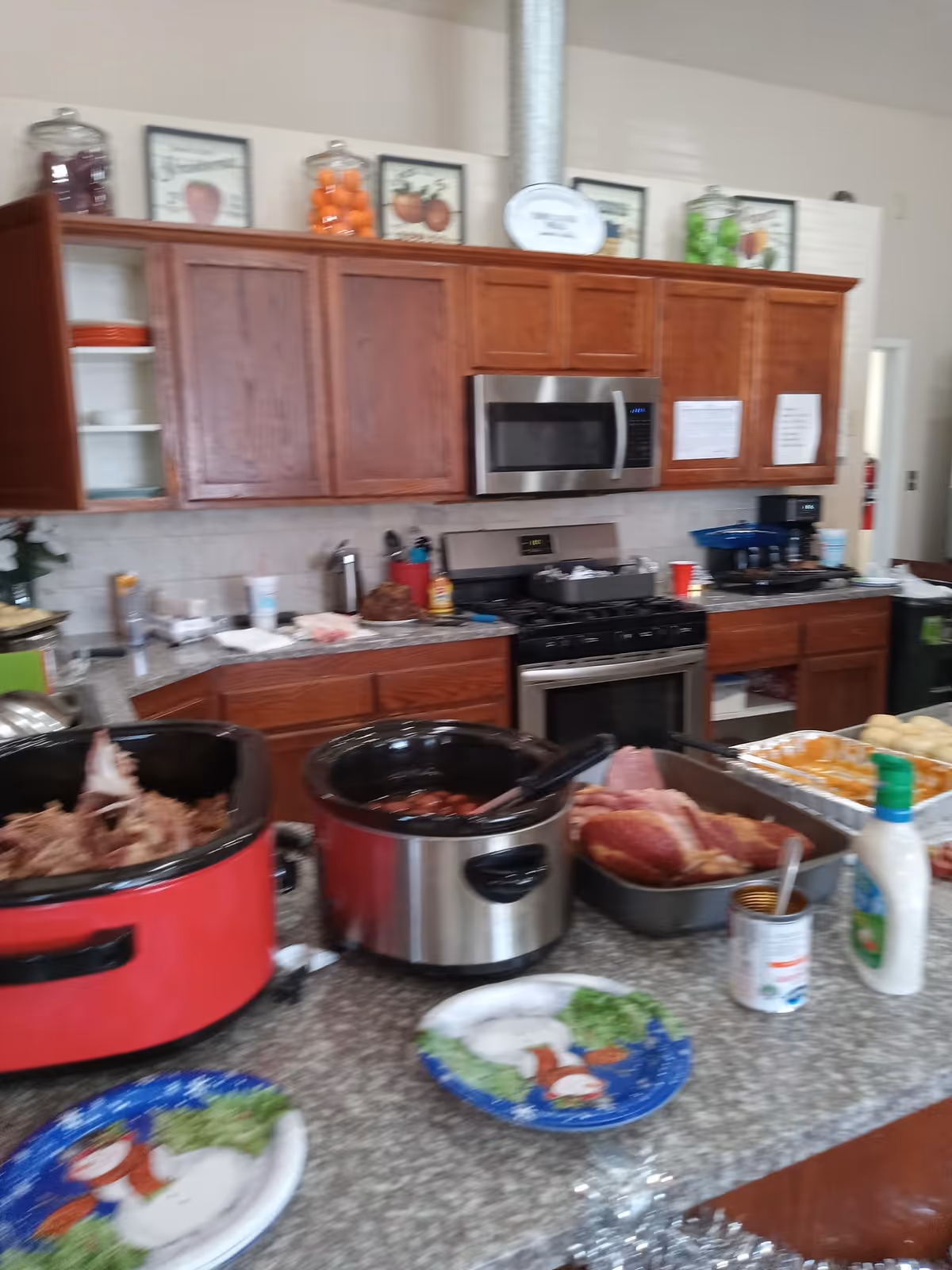 Kitchen countertop with slow cookers, trays of food and paper plates in front of wooden cabinets, a stainless steel microwave and a stove.
