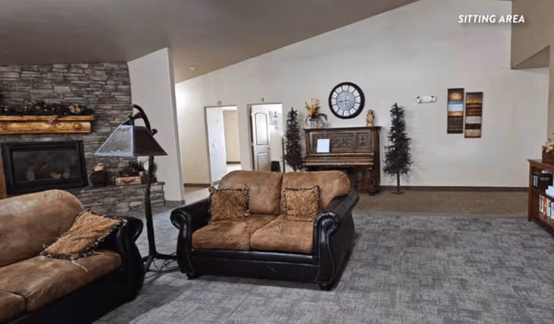 A sitting area in an assisted living facility featuring two brown and black leather sofas with cushions, a stone fireplace with a wooden mantle, a floor lamp, a piano against the wall with decorative items on top, two artificial trees, a large wall clock, and some wall art. The carpeted floor and neutral wall colors create a cozy atmosphere.