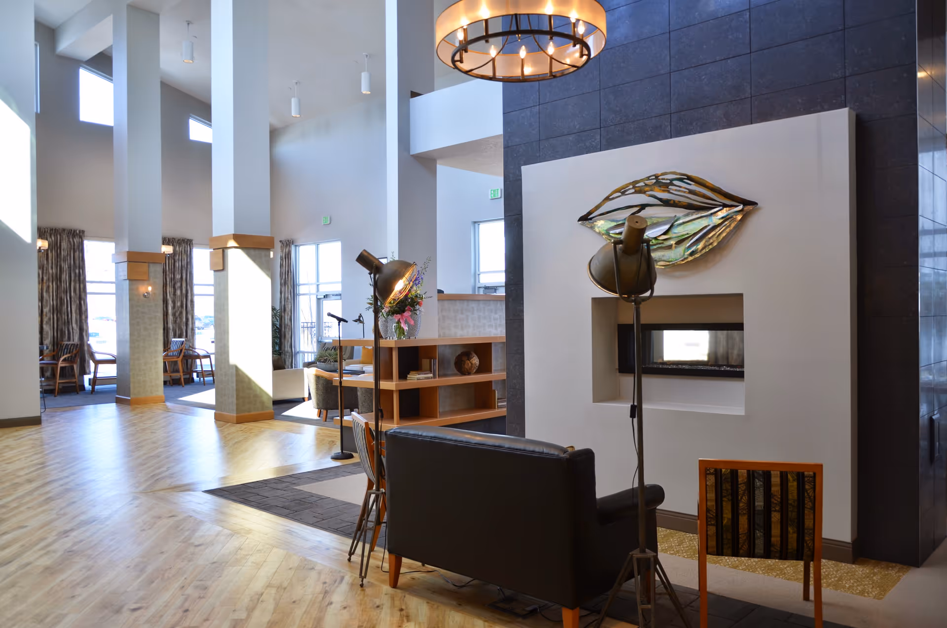 A spacious and modern living area with high ceilings and large windows allowing natural light to fill the room. The space features a black leather sofa, a wooden chair, and two vintage-style floor lamps. A contemporary fireplace is set into a white wall with a decorative metallic art piece above it. The floor is light wood, and there are tall white pillars with wood accents. In the background, there are additional seating areas with tables and chairs near the windows.
