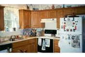 A cozy kitchen with wooden cabinets, a white refrigerator covered with magnets and photos, a white stove with a range hood, a sink under a window with lace curtains, and various kitchen utensils and decorations on the countertops and shelves.