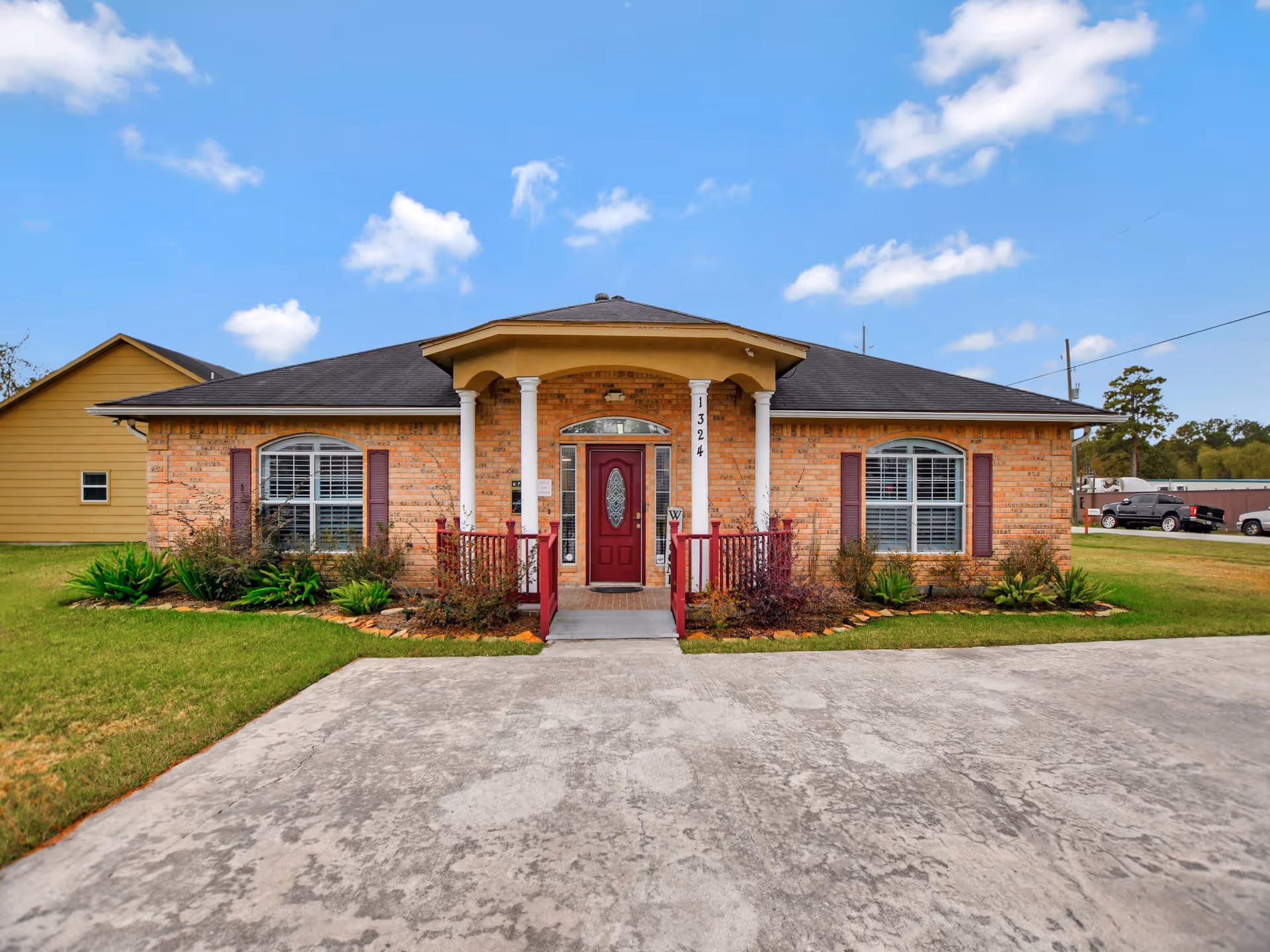 Front exterior view of a single-story brick building with a dark shingled roof, two white columns supporting a small covered porch, a red front door with decorative glass, and two windows with white shutters on either side. The building is surrounded by green grass and landscaped plants, with a concrete driveway leading up to the entrance under a partly cloudy blue sky.