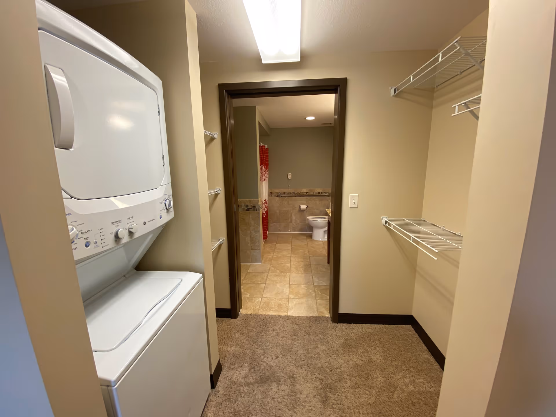 Laundry area with a stacked washer and dryer on the left, beige walls, carpeted floor leading to a bathroom with tiled floor, a toilet, and a shower with a red and white curtain visible in the background. There are wire shelves on the right wall in the laundry area.