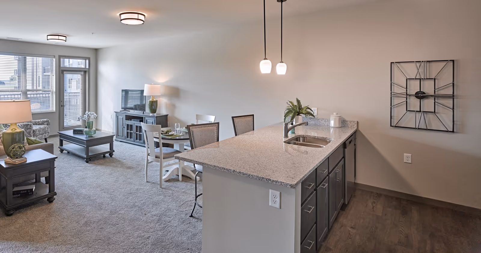 Interior view of a senior living apartment featuring a kitchen island with a sink and two pendant lights, a small dining table with four chairs, a living area with a TV on a stand, a coffee table, an armchair, and a side table with a lamp. Large windows and a glass door lead to a balcony, allowing natural light to fill the space. A decorative wall clock is mounted on the wall near the kitchen.