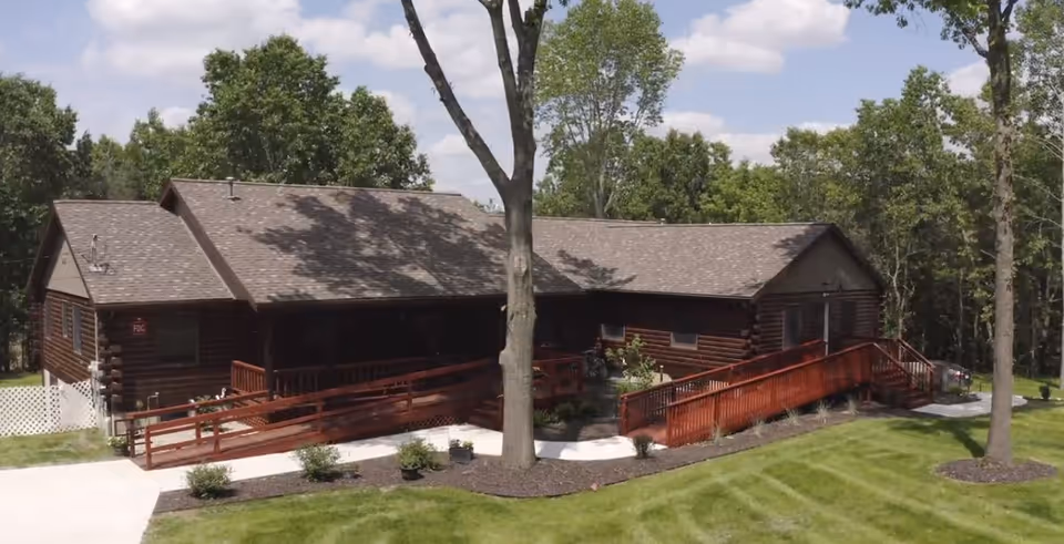 Exterior view of a single-story log cabin style building with a shingled roof, surrounded by green grass and trees under a partly cloudy sky. The building features wooden ramps leading to the entrances on either side.
