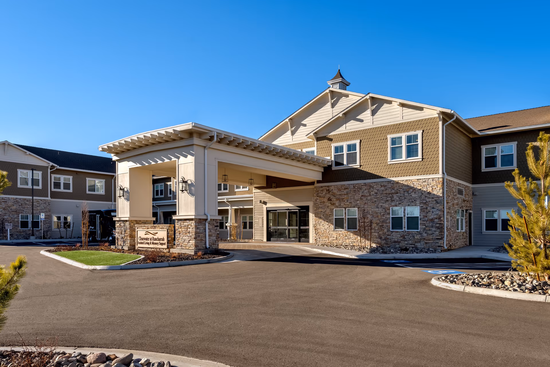Exterior view of Clearwater at Rancharrah, a senior living facility with assisted living and memory support. The building features a covered entrance with stone and beige siding, multiple windows, and a clear blue sky above.