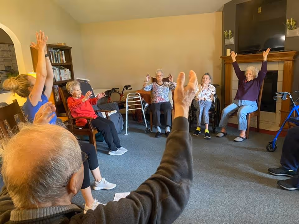 A group of elderly individuals seated in a circle in a living room, participating in a seated exercise or stretching activity with their arms raised. The room has a carpeted floor, a fireplace with a TV above it, and mobility aids like walkers and wheelchairs are visible.