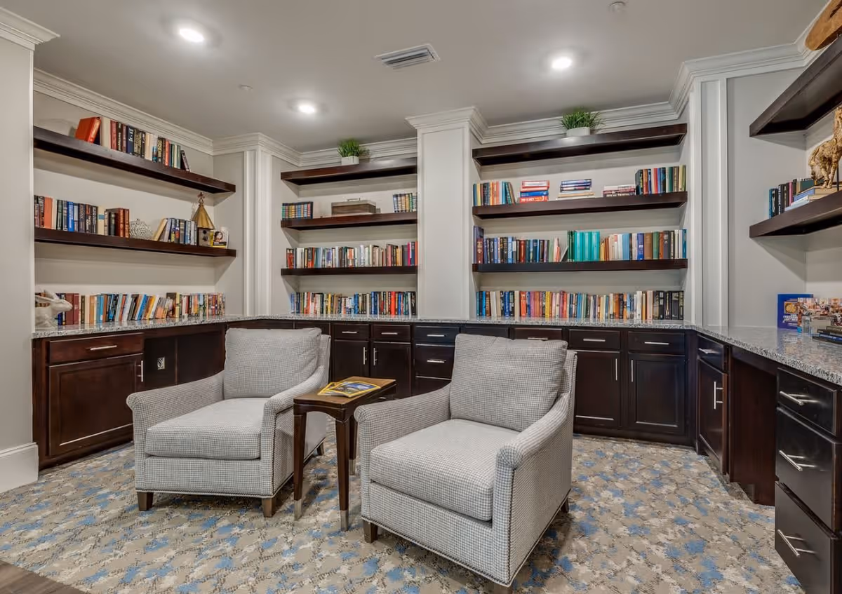 A cozy library reading area featuring two gray armchairs, a small side table, and bookshelves filled with various books. The space has warm lighting and a patterned carpet.