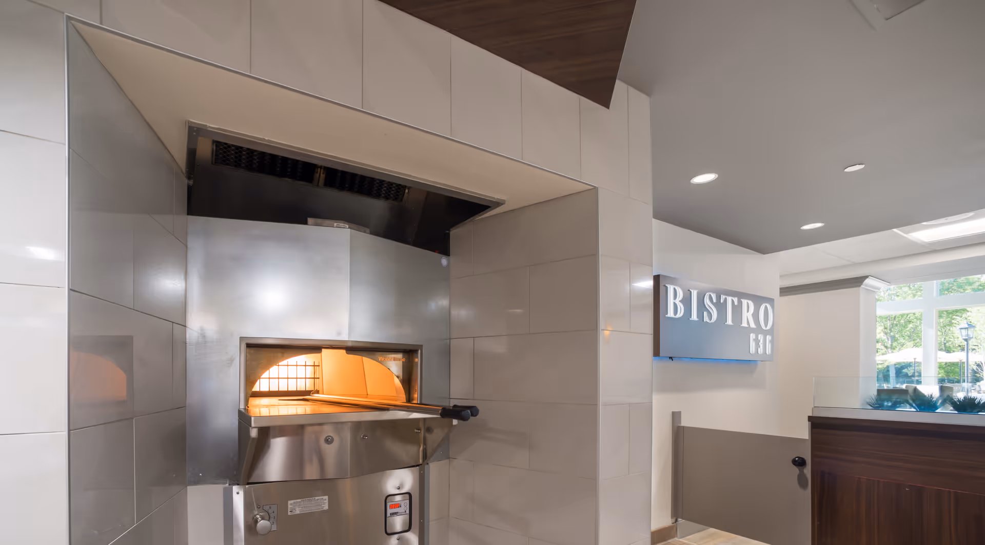 Interior view of a commercial kitchen area with a stainless steel pizza oven built into a tiled wall. To the right, there is a sign that reads 'BISTRO 636' mounted on a white wall, with a window showing greenery outside.
