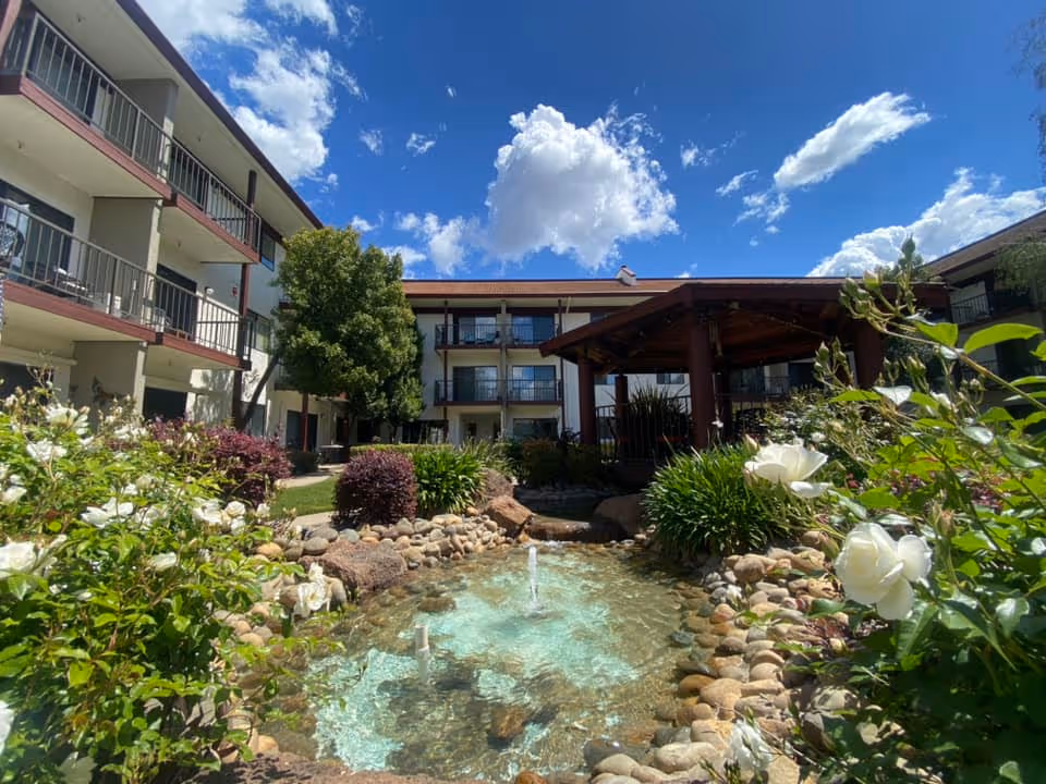 Courtyard of a senior living building with a small fountain surrounded by flowers, a gazebo, and multi-story balconies under a blue sky.