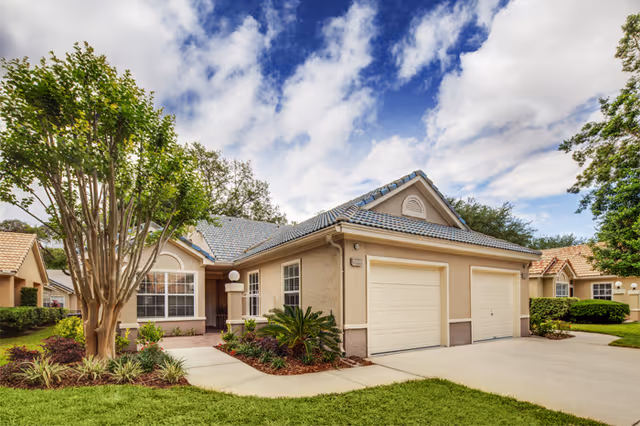 Exterior view of a single-story residential building with a two-car garage, beige walls, and a gray tiled roof. The front yard features a well-maintained lawn, landscaped plants, and a tree. The sky is partly cloudy with patches of blue.