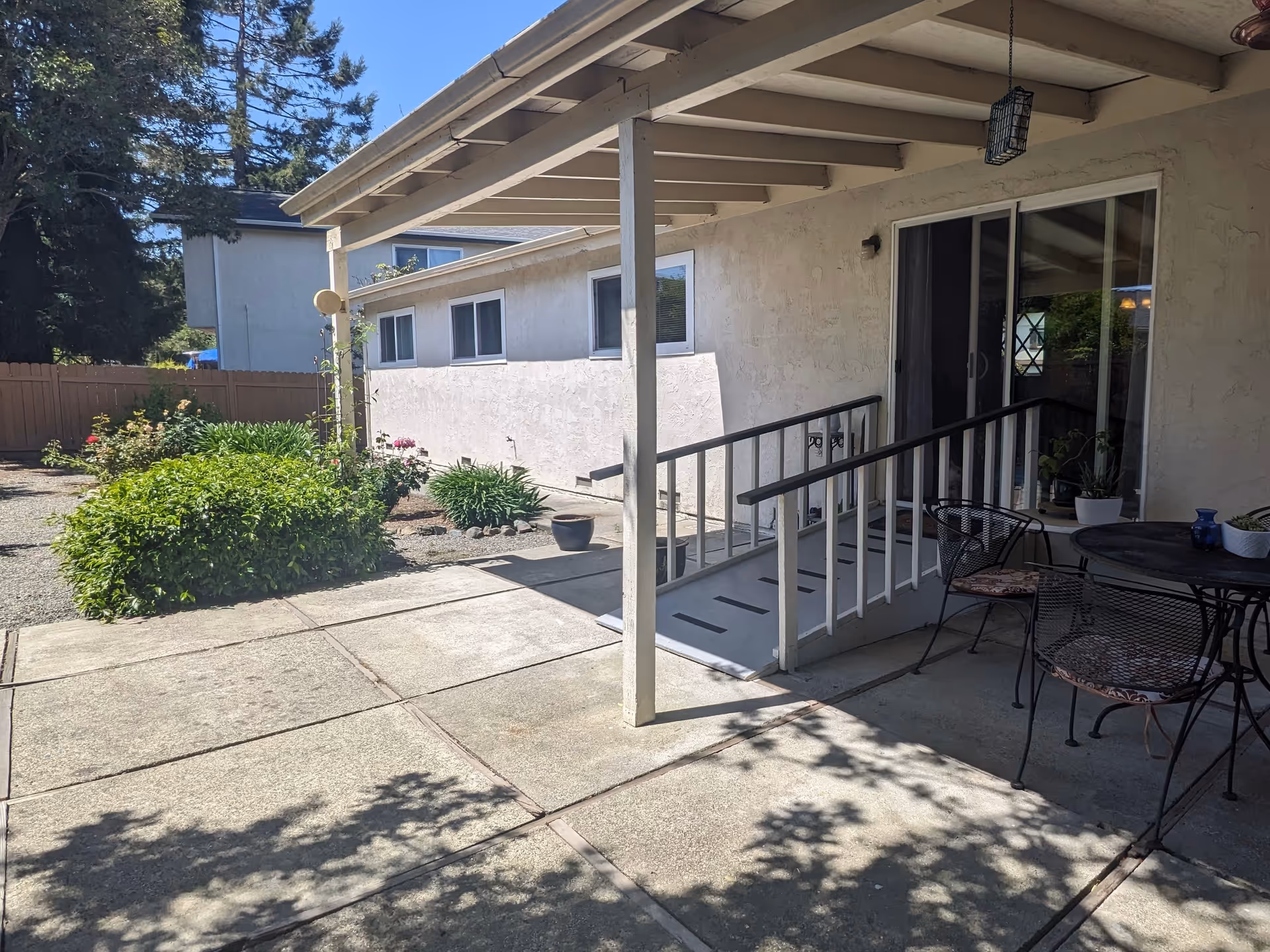 Outdoor patio area with a covered porch featuring a wheelchair ramp and a round metal table with four chairs. There are potted plants on the table and near the sliding glass door. The patio is adjacent to a garden with green bushes and flowers, and a wooden fence is visible in the background.