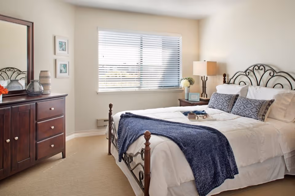 A bright bedroom with a large window covered by white blinds. The room features a bed with white bedding, two patterned pillows, and a dark blue throw blanket. There is a wooden nightstand with a lamp, a small framed picture, and a vase with flowers. On the left side, there is a wooden dresser with a mirror and decorative items on top.
