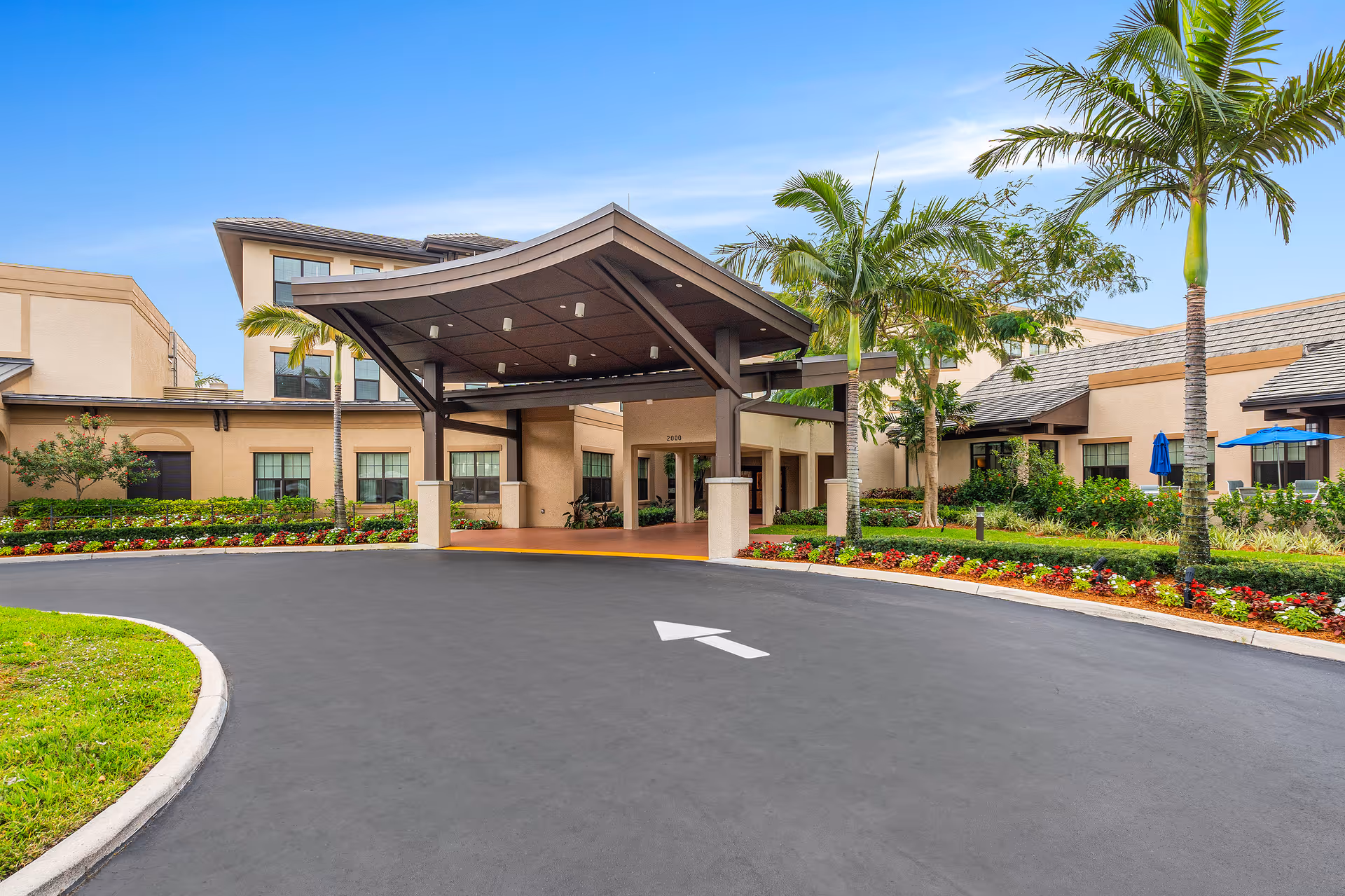 Entrance of a senior living facility with a covered drop-off area, surrounded by landscaped gardens and palm trees under a clear blue sky.