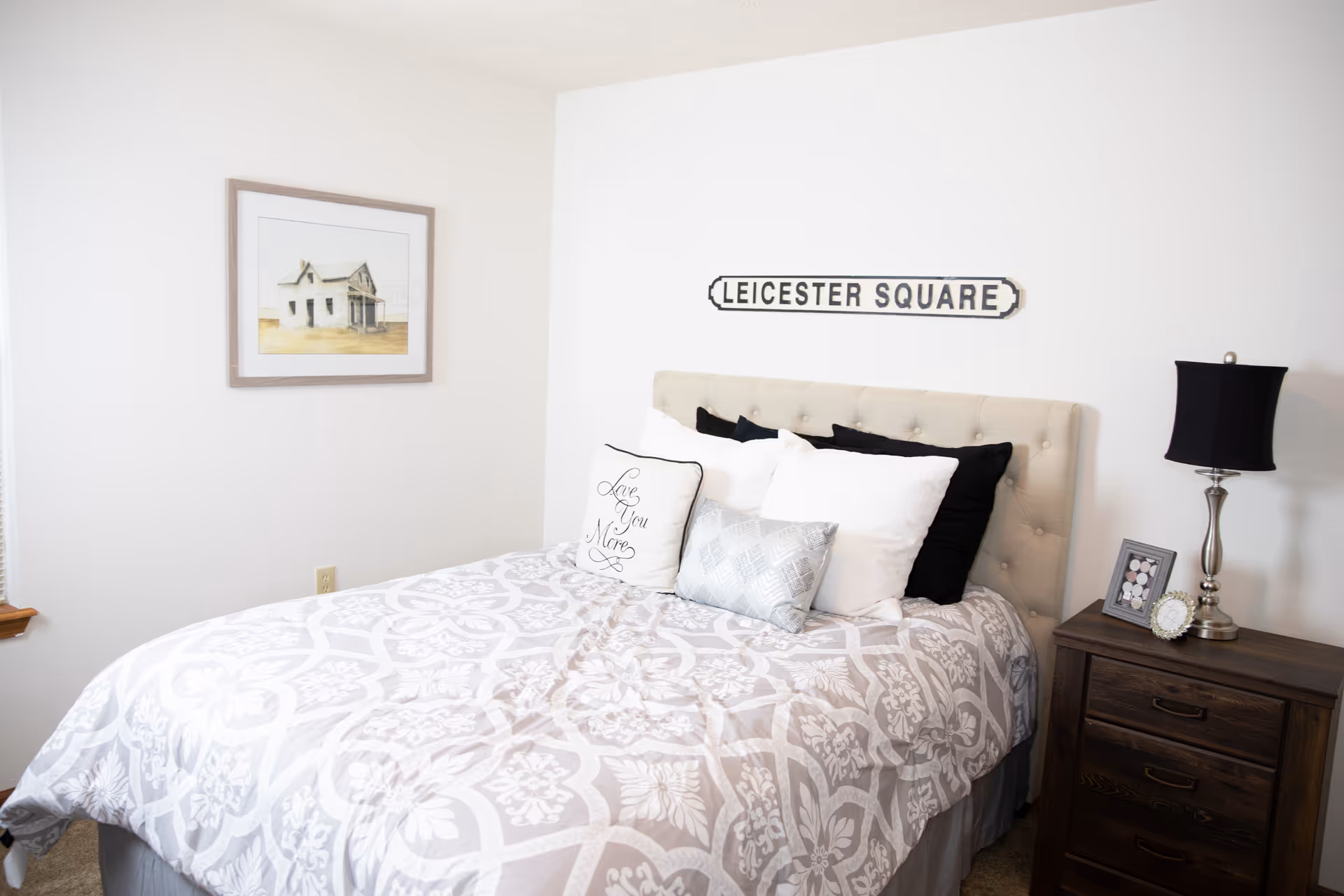 A bedroom with a bed featuring a beige tufted headboard, white and black pillows, and a patterned gray and white comforter. Above the bed is a sign that reads 'LEICESTER SQUARE'. To the right of the bed is a dark wooden nightstand with a silver lamp and two framed pictures. On the left wall is a framed painting of a house.