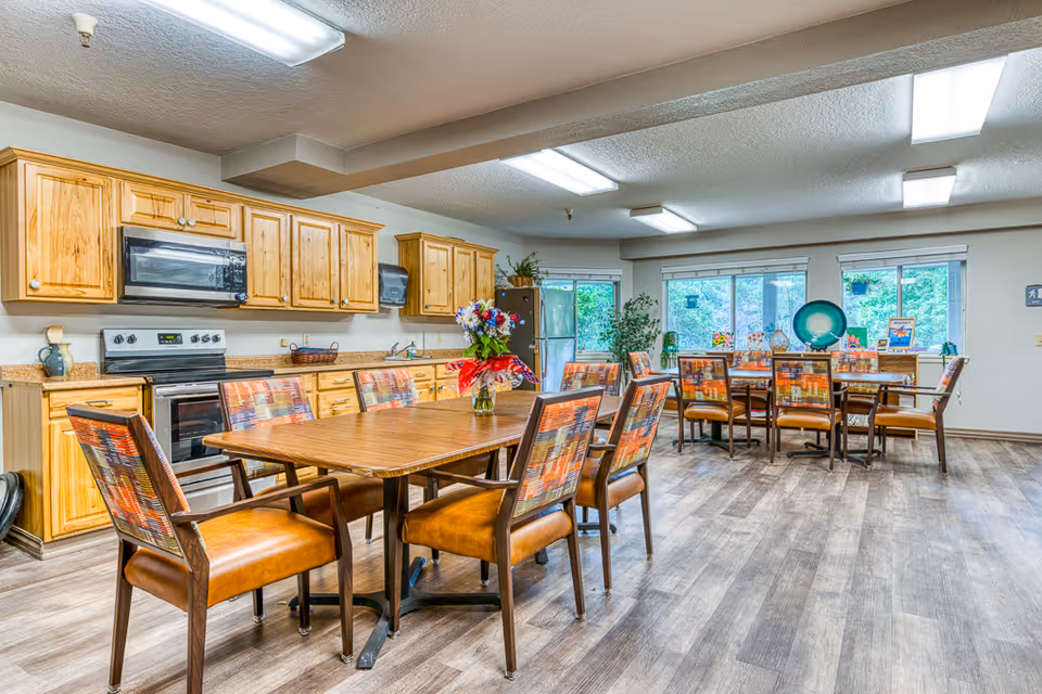 A bright and spacious dining area in a senior living facility featuring wooden tables with colorful cushioned chairs. The room includes a kitchen area with wooden cabinets, a stove, microwave, and refrigerator. Large windows let in natural light and there are decorative plants and flowers on the tables and windowsills.