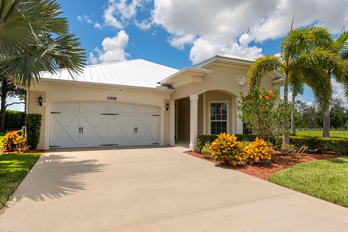 Front exterior of a single-story house with a double garage, driveway, palm trees, and landscaped yard under a blue sky.