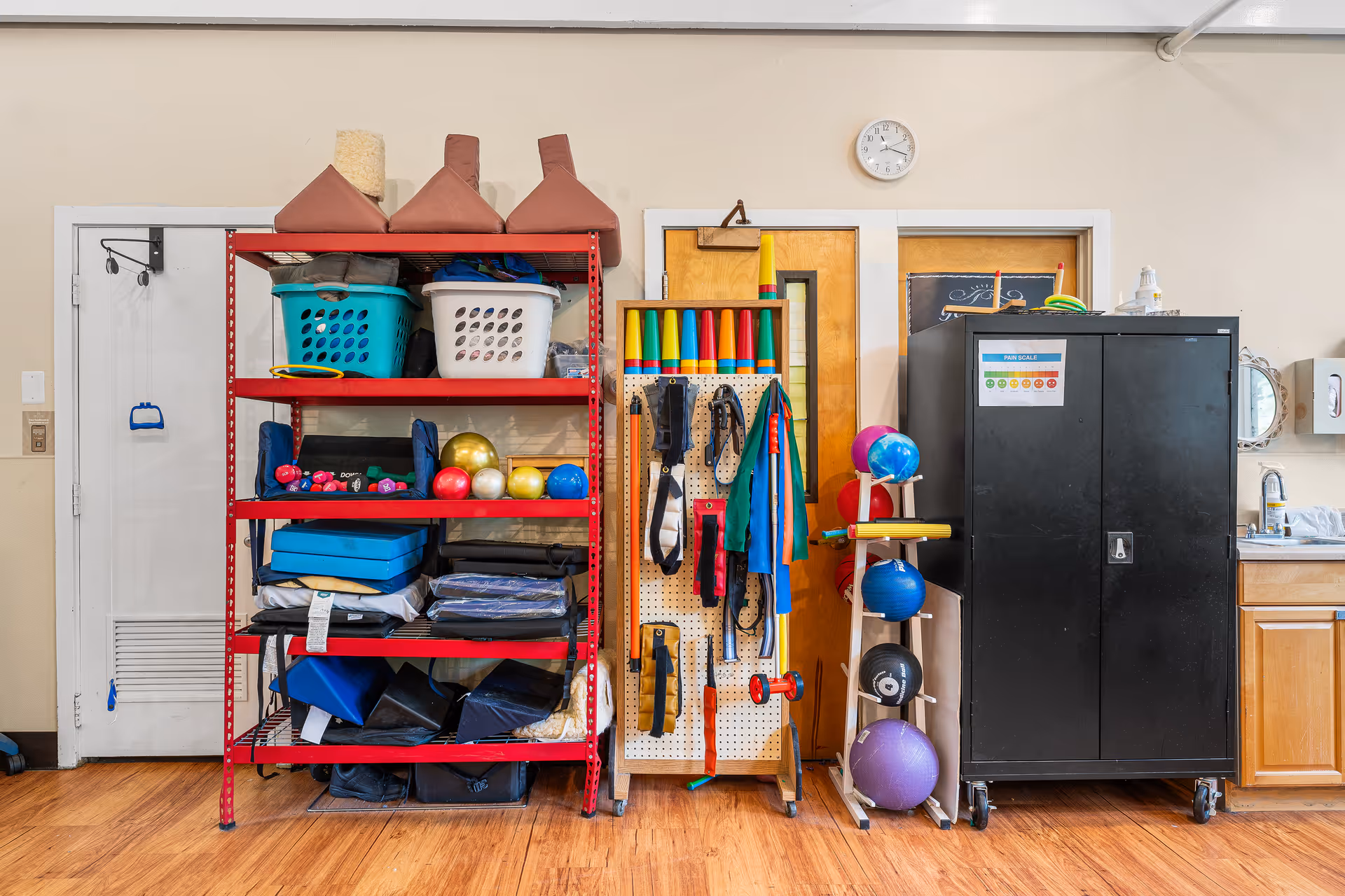 A storage area in a rehab facility with a red metal shelving unit holding exercise balls, weights, mats, and baskets. Next to it is a pegboard with various therapy and exercise equipment hanging, and a rack with more exercise balls. A black cabinet and a wooden cabinet with a sink are also visible. The floor is wooden and the walls are beige with two wooden doors and a clock above them.