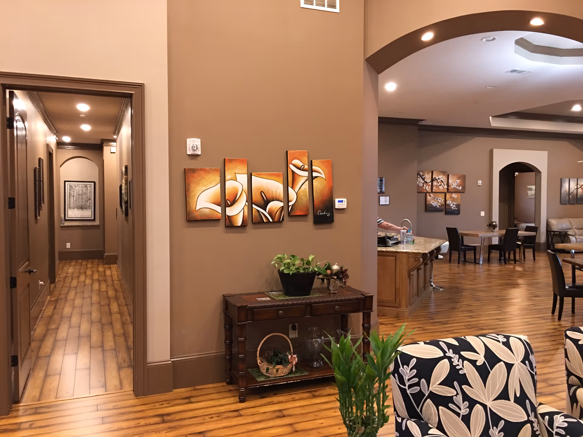 Interior view of a senior living community featuring a hallway with framed artwork, a small wooden table with plants and decorative items, and an open living area with tables, chairs, and additional artwork on the walls. The flooring is wood, and the walls are painted in warm neutral tones.