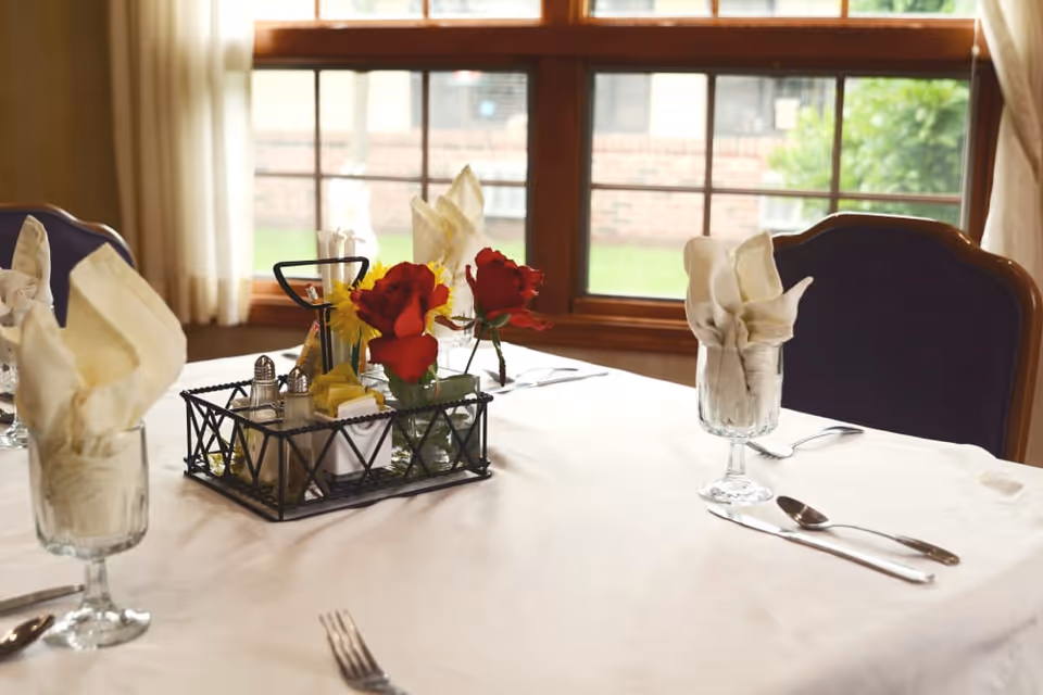 A dining table set with white tablecloth, folded cream-colored napkins in glass goblets, silverware, and a centerpiece featuring red roses and condiments in a black metal holder. The table is positioned near a window with sheer curtains and a view of a brick wall and greenery outside.