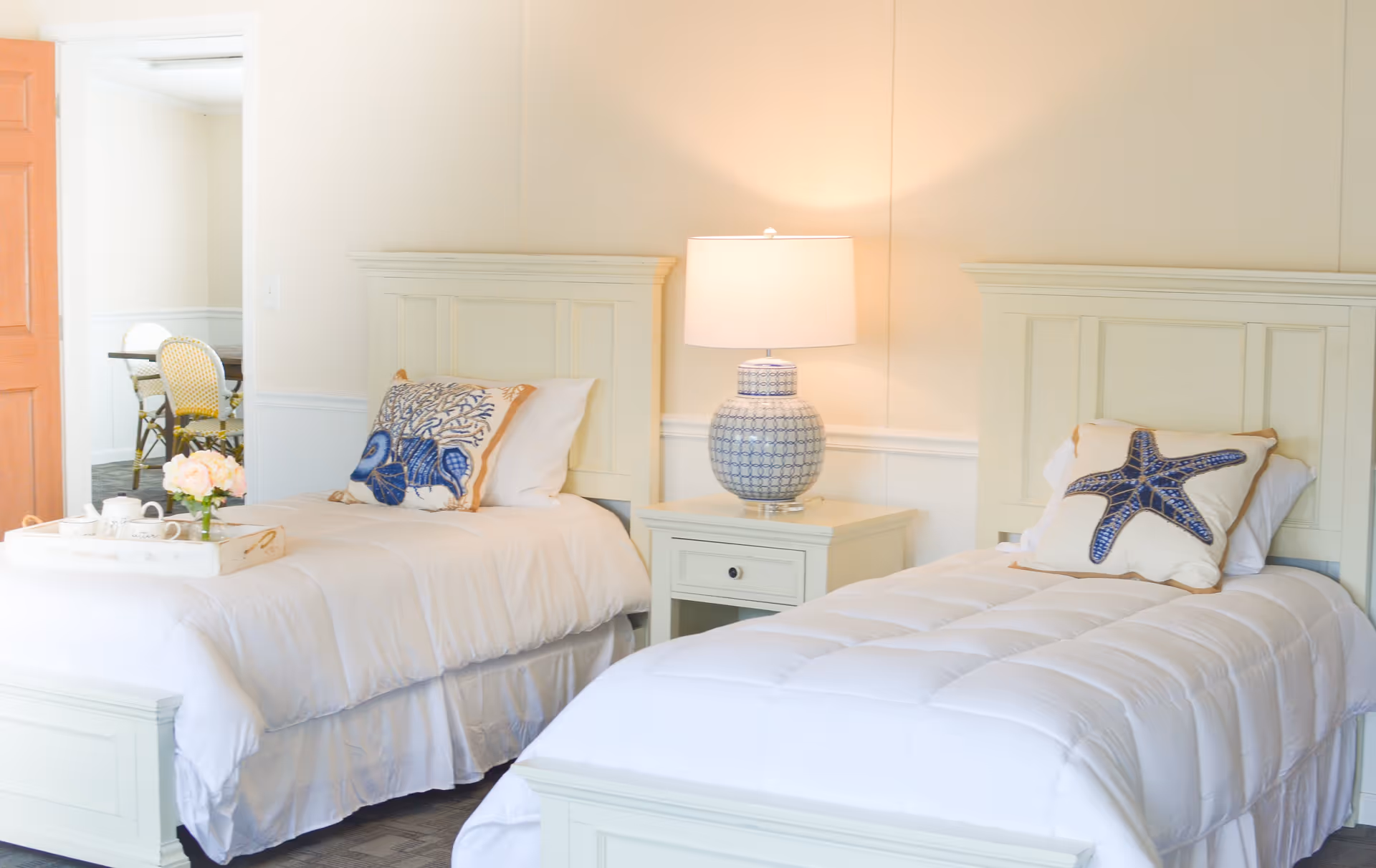 A bright bedroom with two single beds featuring white bedding and decorative pillows with blue sea-themed designs. A nightstand with a patterned lamp is placed between the beds. In the background, there is a glimpse of a dining area with chairs and a table.