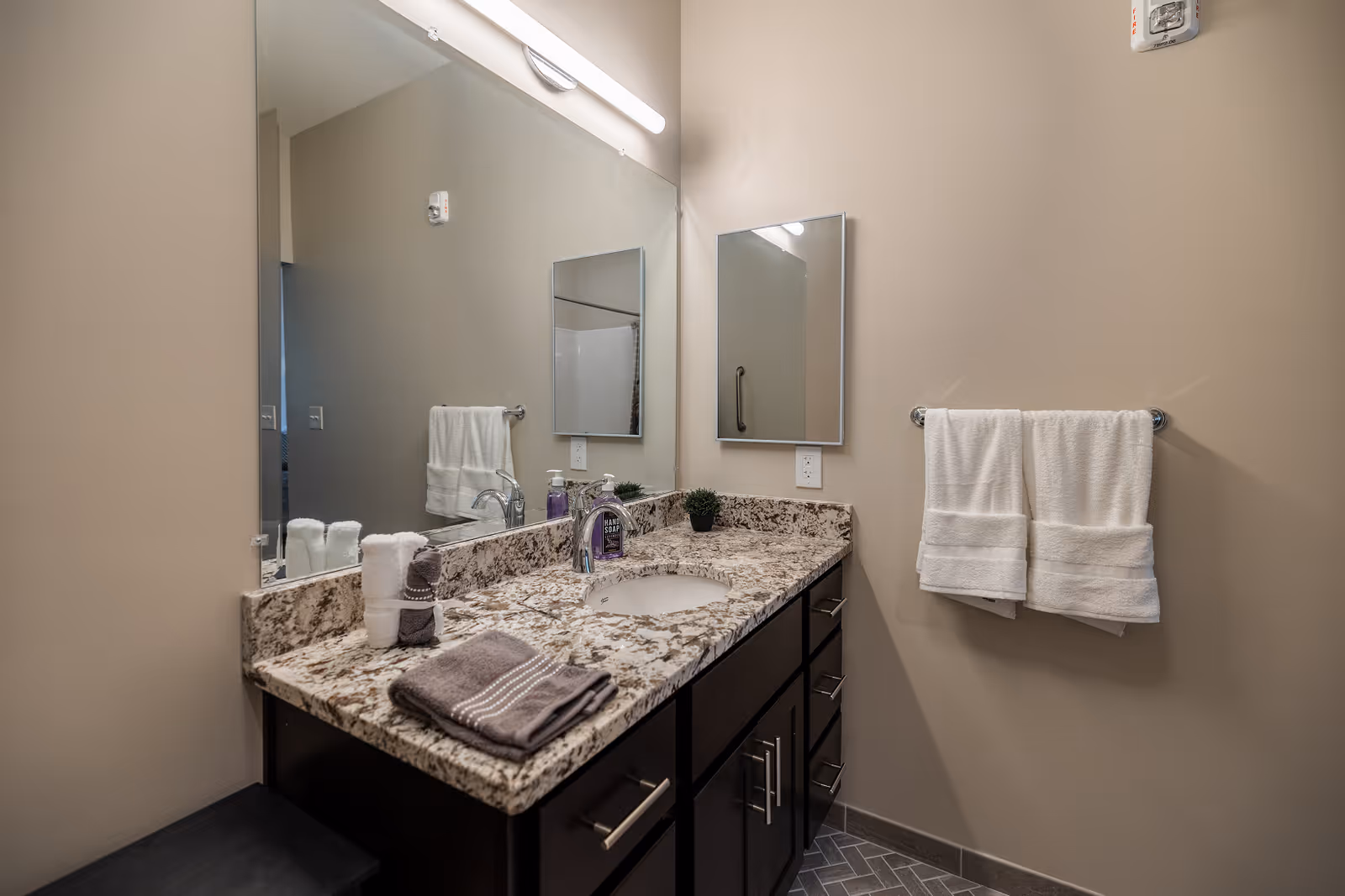 A modern bathroom vanity with a granite countertop, an undermount sink, a chrome faucet, and a large mirror above. On the countertop are neatly folded towels, a small potted plant, and a bottle of hand soap. Two white towels hang on a towel rack mounted on the beige wall to the right. The floor has a herringbone tile pattern.