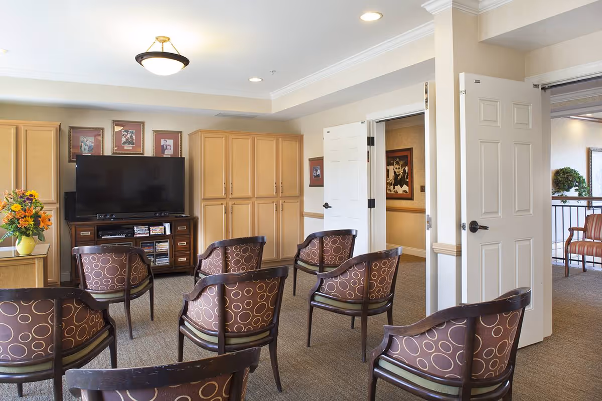 A cozy common room with several patterned armchairs arranged facing a large flat-screen TV on a wooden stand. The room has light-colored walls, wooden cabinets, framed pictures above the TV, and a vase with flowers on a side table. Two white double doors are open, leading to another area with additional seating.