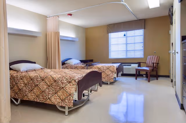 A nursing facility room with two hospital-style beds separated by a privacy curtain. Each bed has a patterned bedspread and a pillow. There is a window with blinds, a wooden chair, and a small table near the window. The room has beige walls and a clean, polished floor.