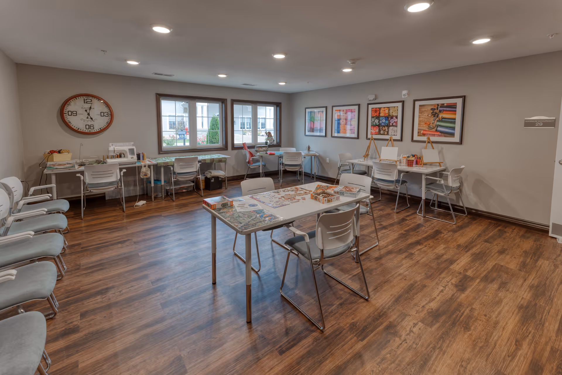 A well-lit activity room with wooden flooring and light gray walls. The room contains several white chairs arranged along the left wall and around tables in the center and right side of the room. On the tables are puzzles, art supplies, and sewing materials. The walls are decorated with framed pictures of colorful yarn and sewing tools. Large windows provide natural light and a view of the outside.