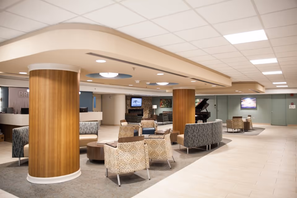 A spacious and well-lit interior common area with modern furniture including patterned armchairs and sofas arranged around circular wooden tables. The room features large wooden columns, a tiled floor, and a ceiling with recessed lighting. In the background, there is a reception desk, a fireplace with a TV mounted above it, and a grand piano. The walls are decorated with artwork and the overall atmosphere is welcoming and comfortable.