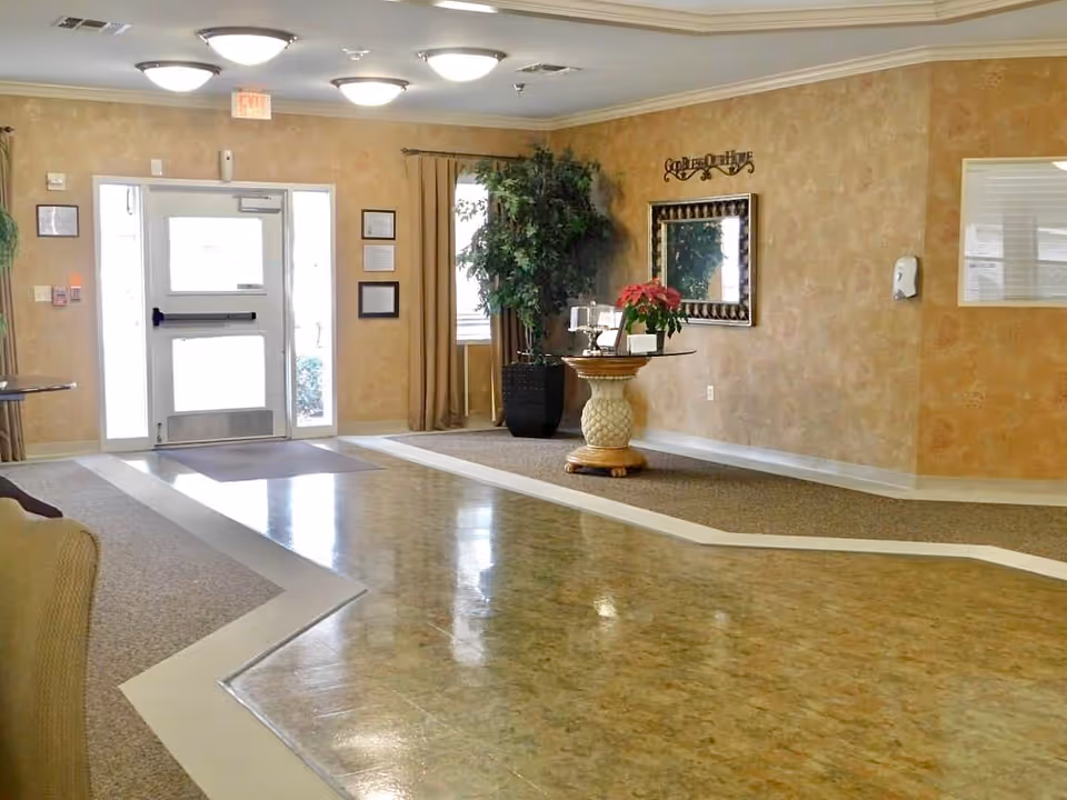 Spacious nursing home lobby with a decorative round table and poinsettia, potted plant, mirror on the wall, and a glass exit door.