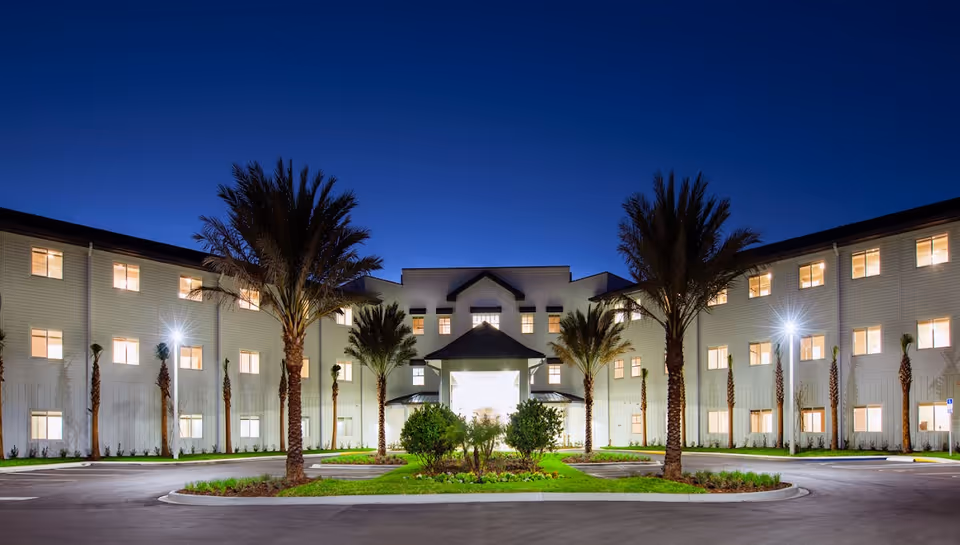 Night view of the front exterior of a large, well-lit assisted living and memory care facility named Paradise Pointe. The building is three stories tall with numerous windows illuminated from inside. There are palm trees and landscaped greenery in front of the entrance, with a circular driveway leading up to the main entrance.