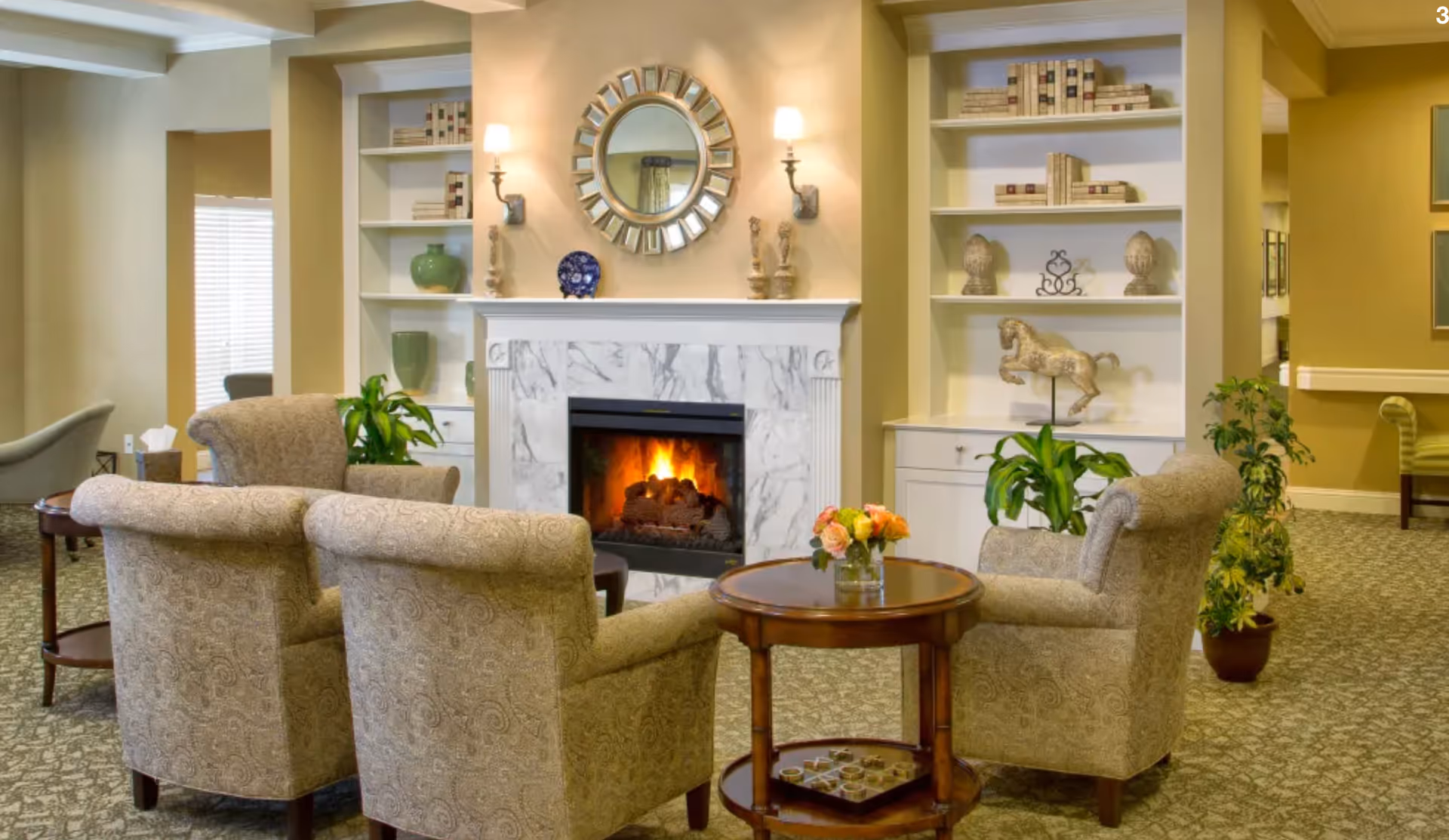 A cozy living room area with four upholstered armchairs arranged around a small round wooden table with a vase of flowers. In the background, there is a white marble fireplace with a fire burning, flanked by built-in shelves decorated with books, vases, and sculptures. The room has warm lighting and a patterned carpet.