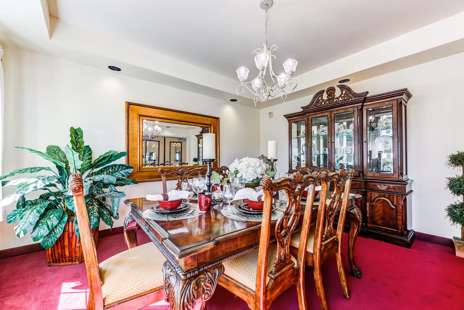 A bright dining room with a polished wooden dining table set for six, featuring red bowls, wine glasses, and white napkins. The room has a large wooden china cabinet filled with glassware and decorative items, a large mirror on the wall, a chandelier hanging from the ceiling, and potted green plants in the corners. The floor is covered with red carpet.
