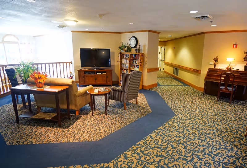 A cozy common area in a senior living facility featuring a TV on a wooden stand, a bookshelf, a beige armchair, a striped armchair, a small round table, and a wooden table with a flower arrangement. The area has patterned carpet with a blue border and a piano with a chair against the wall in the background.
