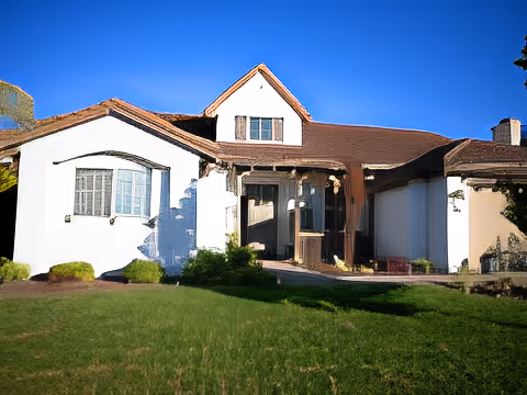 Front exterior view of a single-story house with white walls, brown roof, and a small porch area. The house is surrounded by a green lawn and bushes under a clear blue sky.