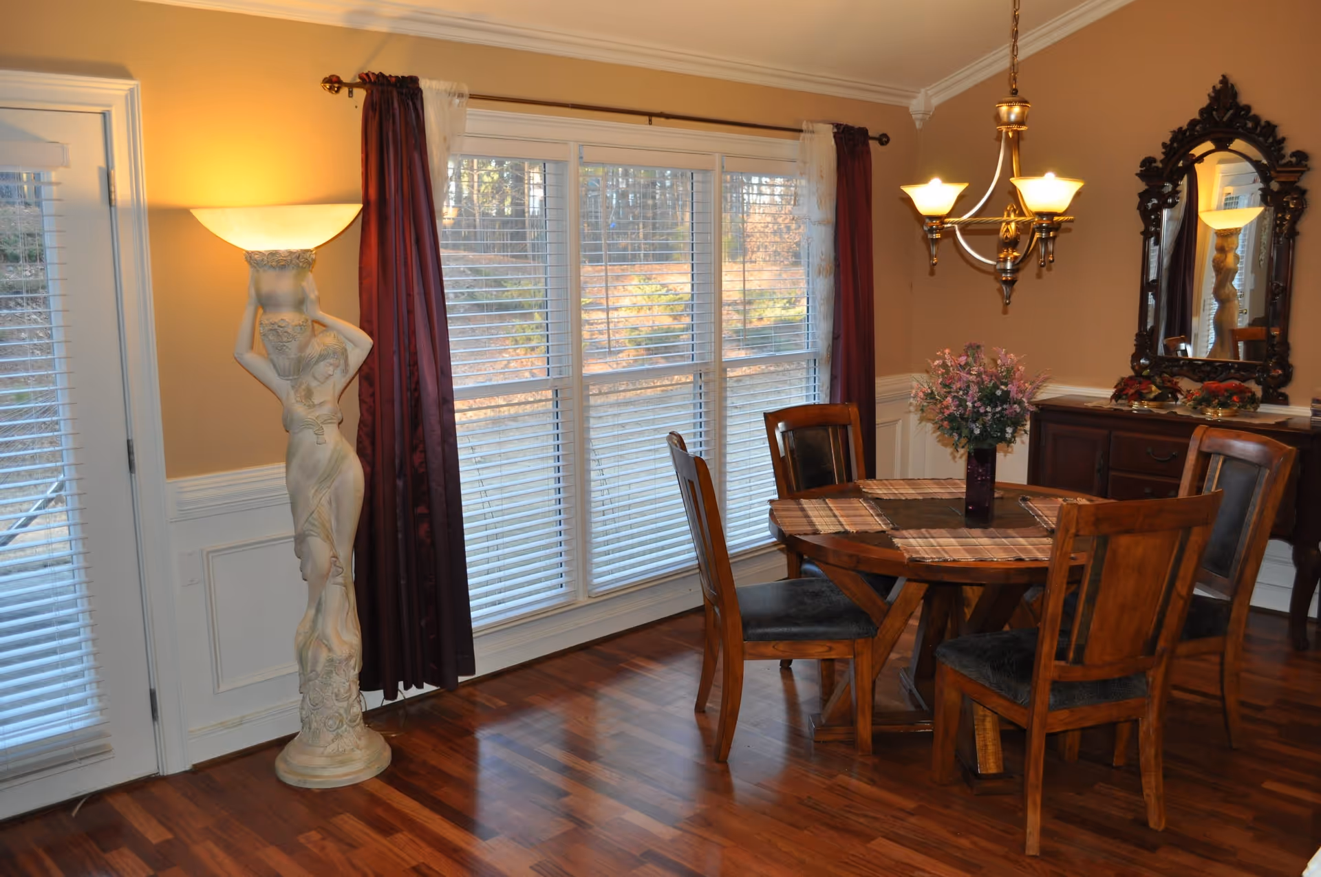 Wooden dining room featuring a round table with four chairs and a floral centerpiece, a large window with blinds and curtains, and decorative floor lamp and chandelier.
