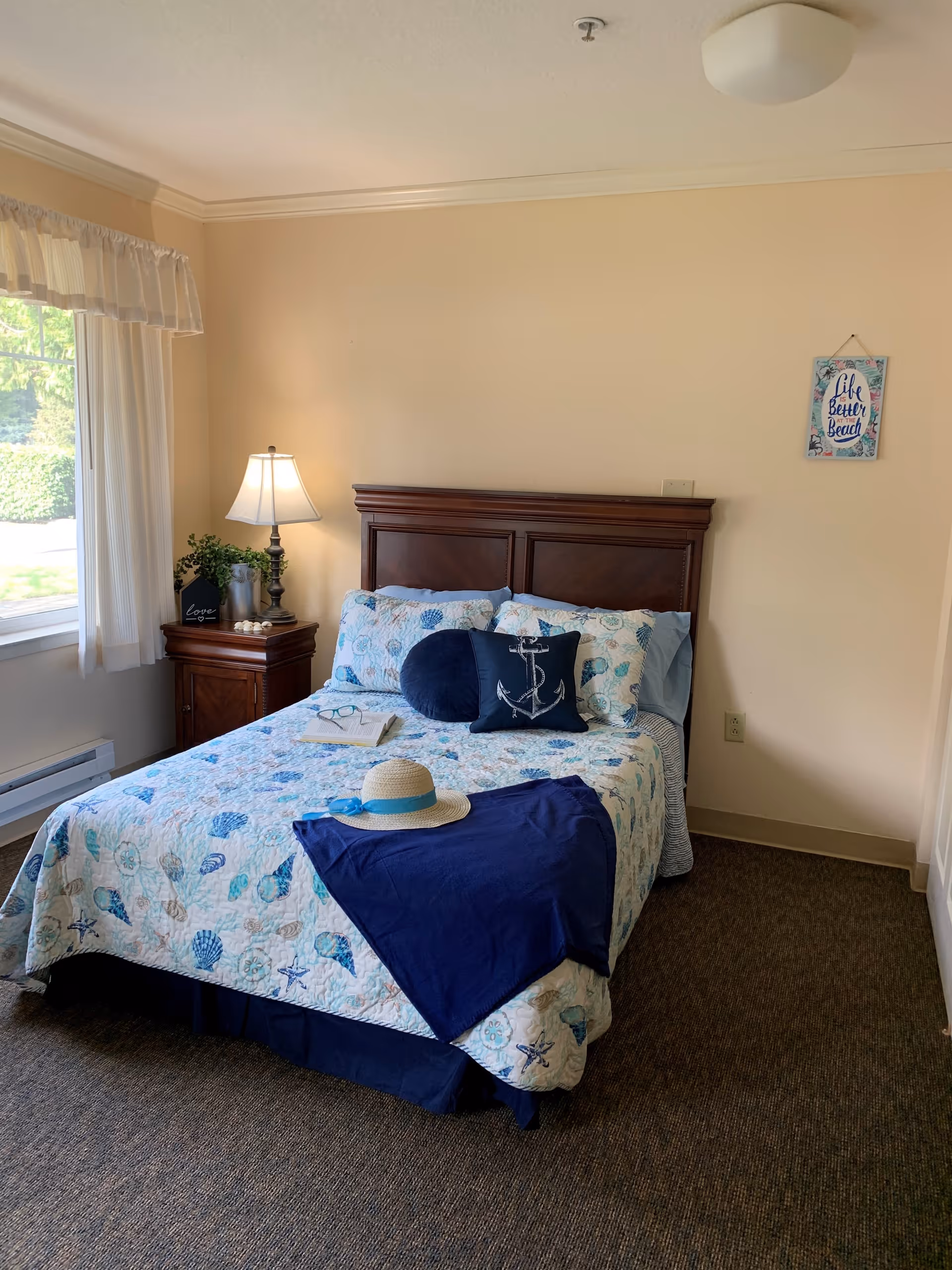 A cozy bedroom with a bed featuring a seashell-patterned quilt and decorative pillows, including one with an anchor design. A navy blue throw and a straw hat with a blue ribbon are placed on the bed. Next to the bed is a wooden nightstand with a lamp, a small plant, and decorative items. A window with sheer white curtains lets in natural light. A wall decoration with the phrase 'Life is Better at the Beach' hangs on the beige wall.