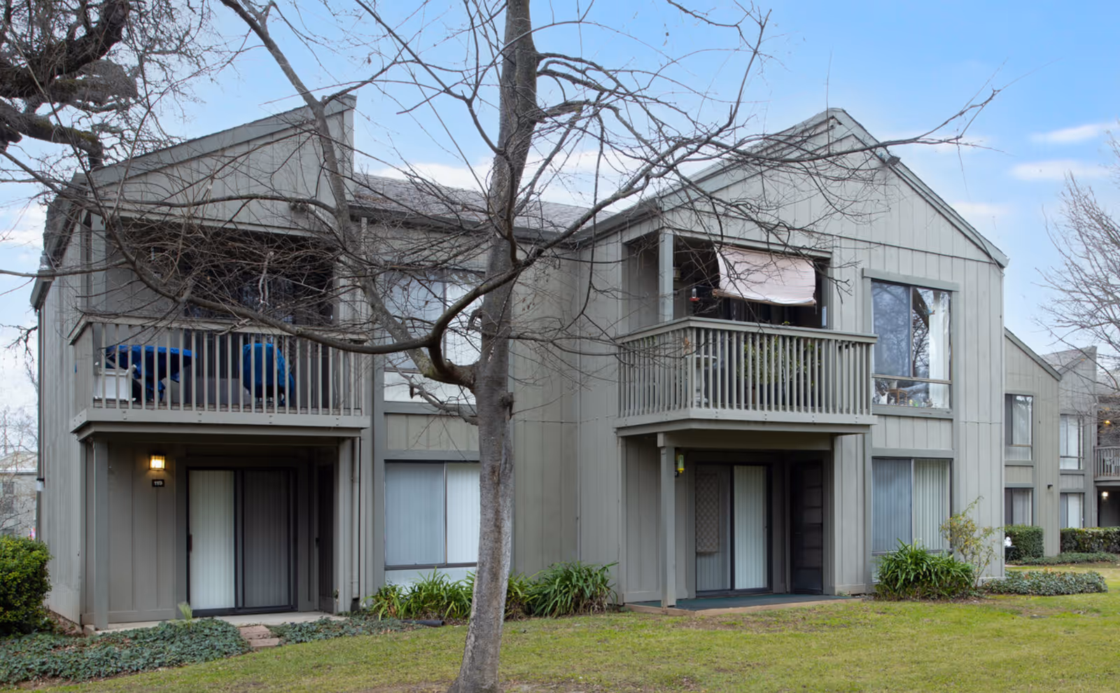 Exterior front of a two-story gray residential building with second-floor balconies, ground-level patios, and a leafless tree on a grassy lawn.