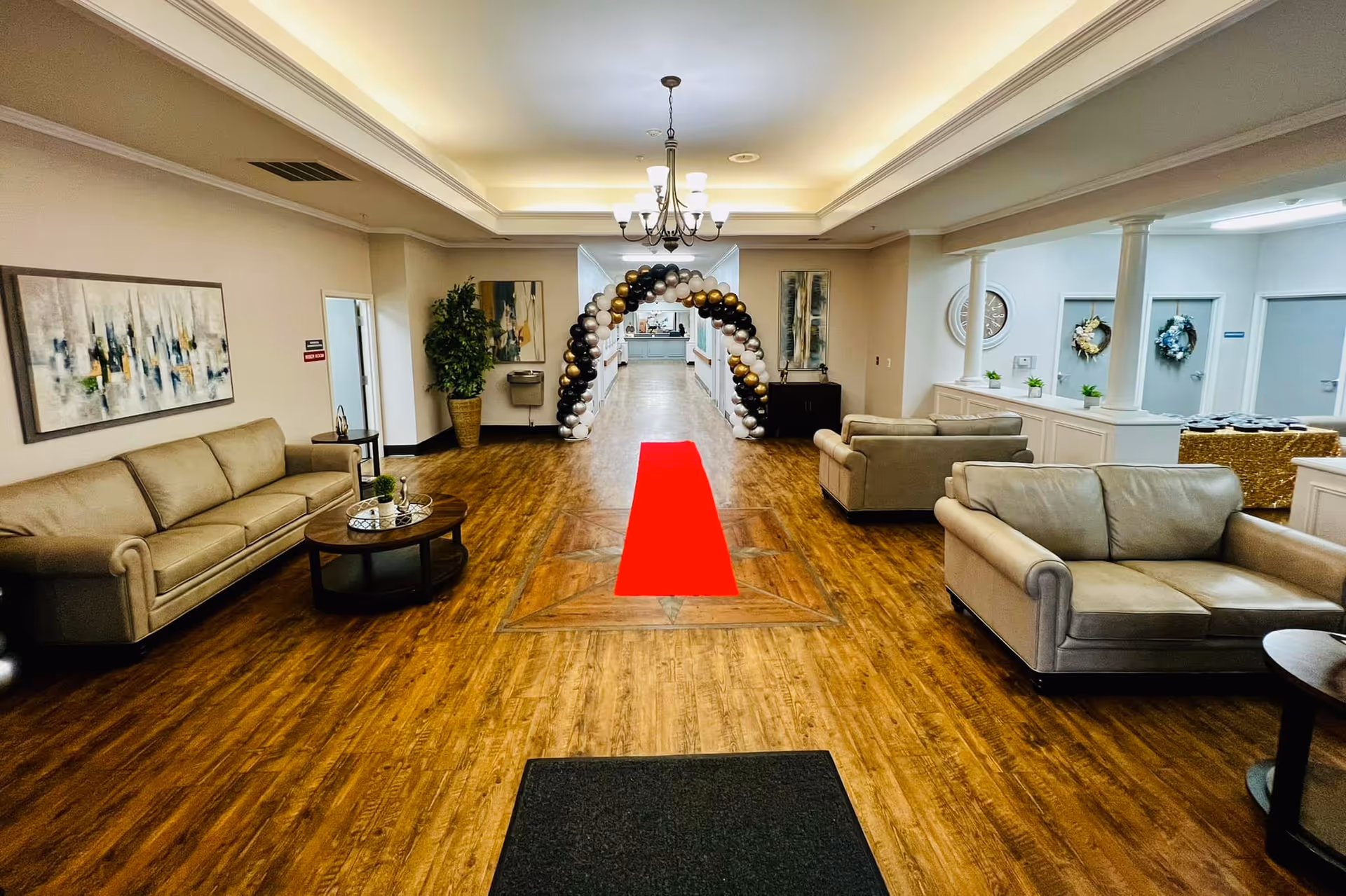 A reception-style interior with sofas and tables flanking a wood-floor aisle topped by a red carpet runner and a gold-and-white balloon arch.