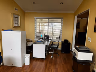 Small office room with desks and rolling chairs, a printer and filing cabinet, wood floor and a window with blinds.