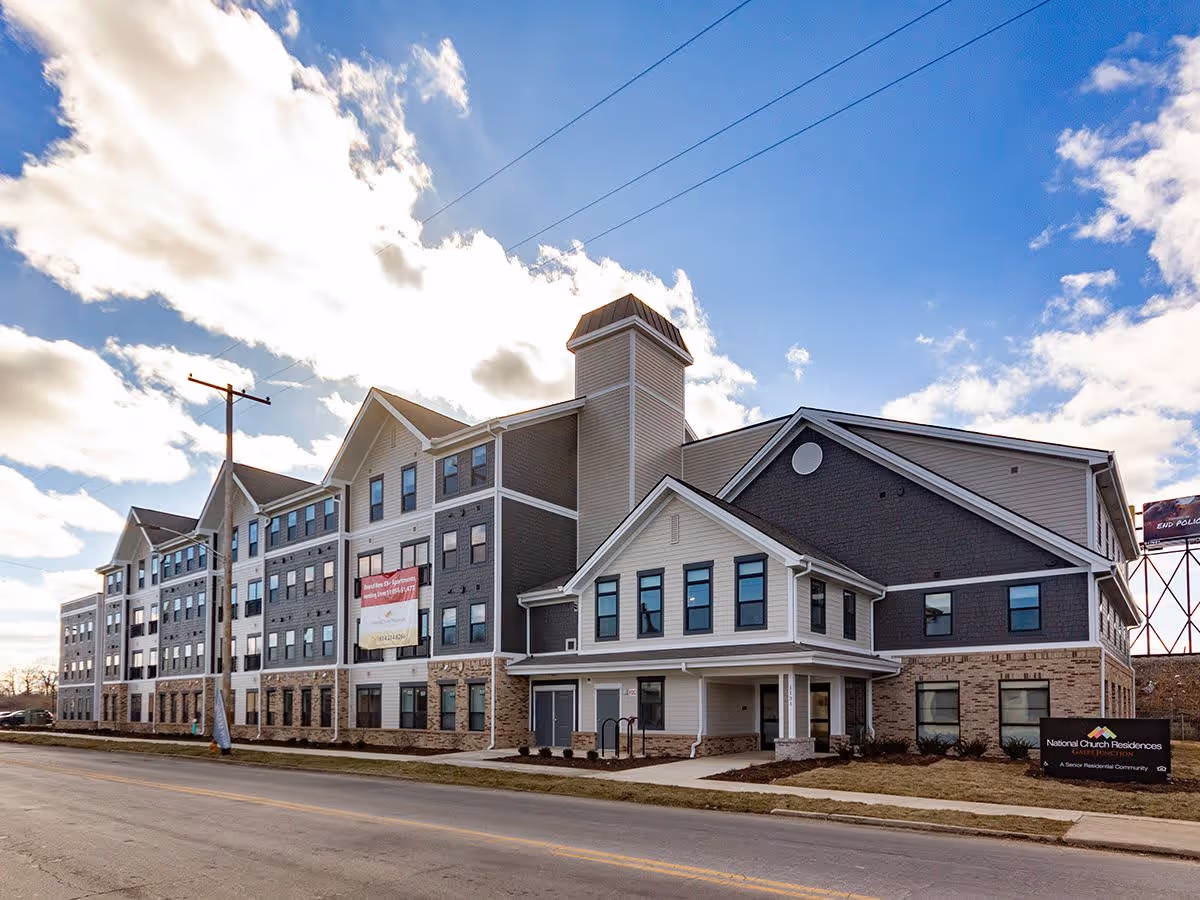 Front exterior of a large multi-story senior living building with pitched roofs and a brick-and-siding facade under a blue sky.