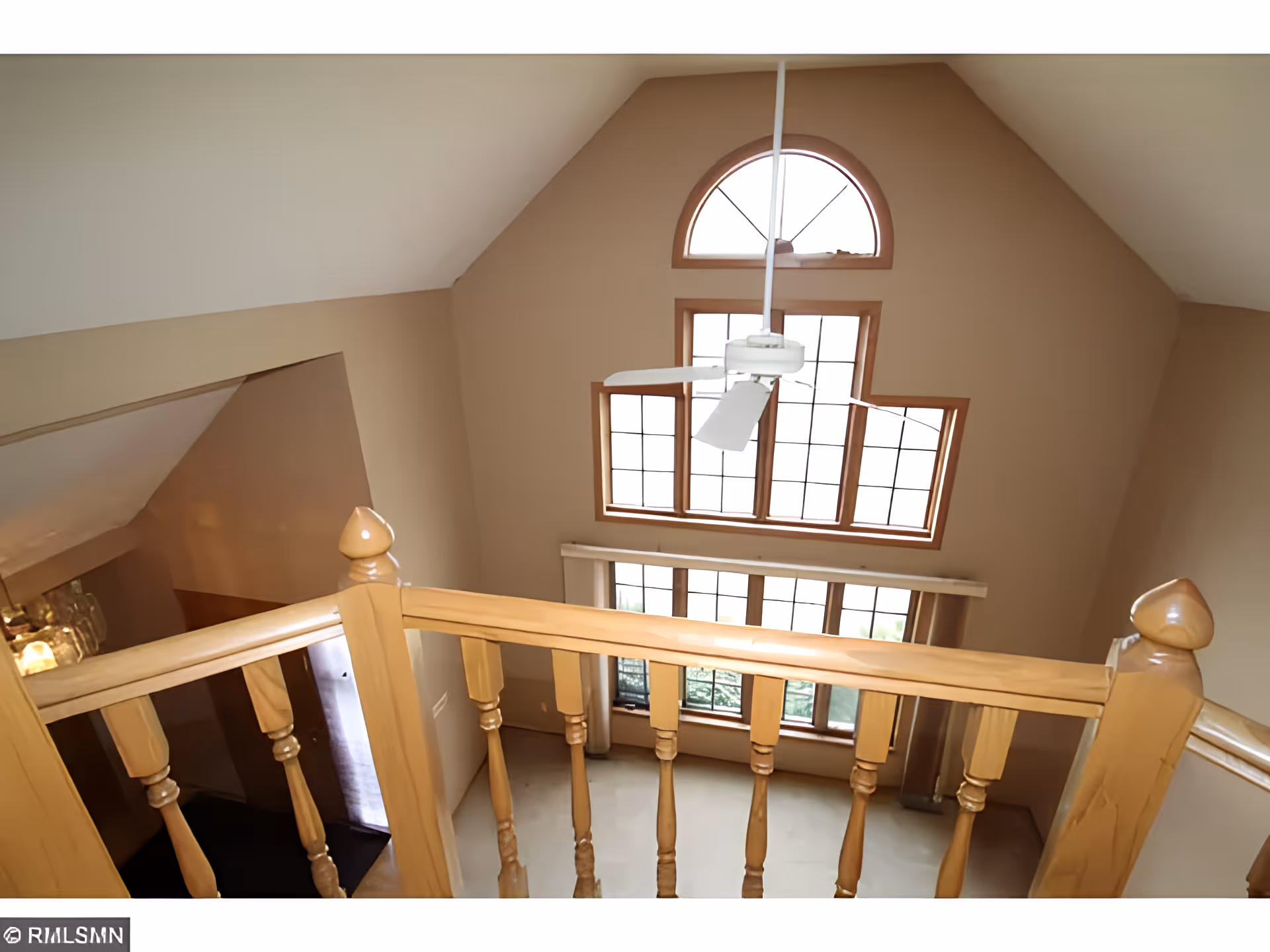 View from an upper floor looking down into a living area with large windows featuring a semi-circular window at the top. The room has beige walls, a ceiling fan, and wooden railing in the foreground.