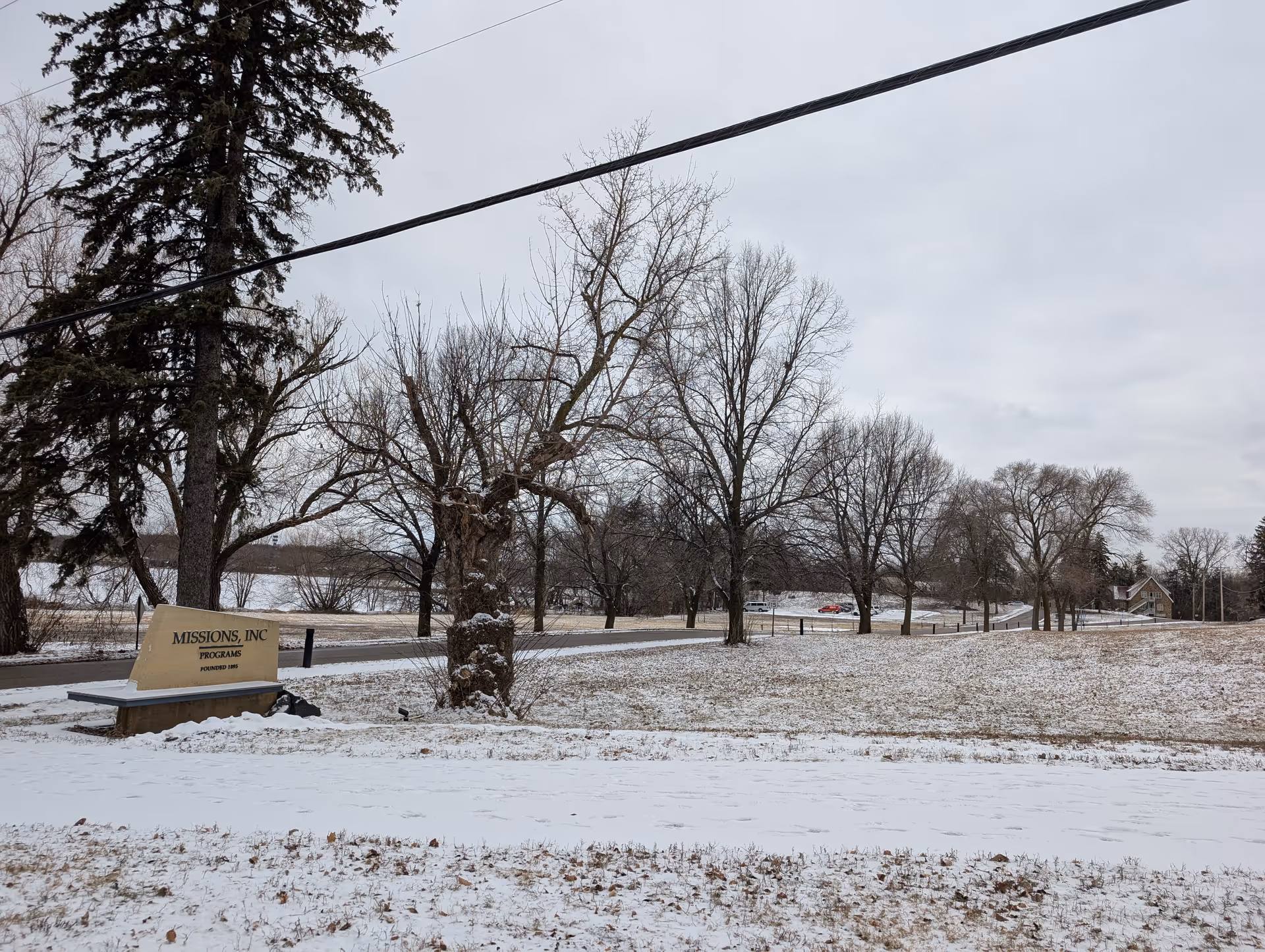 Snow-covered landscape with leafless trees and a road running through it. A sign near the road reads 'MISSIONS, INC PROGRAMS FOUNDED 1883'. The sky is overcast and there are a few parked cars visible in the distance.