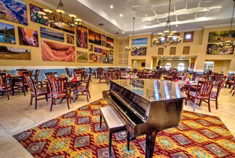 Spacious dining room with a grand piano in the foreground, many tables and chairs, and colorful landscape artwork on the walls.